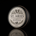 A round metal tin of St. Aries Botanical Apothecary Beard Balm, featuring a detailed black and white label with a ram's head and botanical illustrations, set against a solid black background with a reflection below.